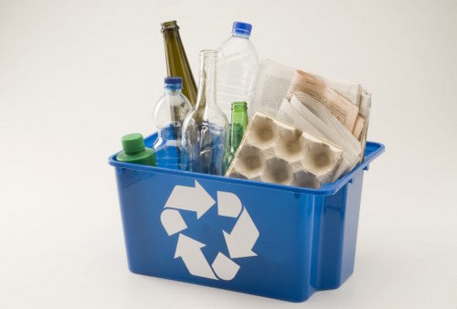 A blue recycling bin filled with various types of rubbish, including clear and green plastic bottles, an empty glass bottle, a pile of crumpled and stained newspapers, and a plastic egg carton. The bottles are of different shapes and sizes, with some having blue and green caps, and the empty glass bottle standing upright among the plastic items. The newspapers are stacked and slightly spilling over the edge of the bin, exhibiting creases and discoloration. The plastic egg carton appears intact and is placed toward the front of the bin. The background is plain and light-colored, emphasizing the contents of the bin, which is situated on a neutral grey or beige surface. The bin bears a white recycling symbol on its side, indicating its purpose for recyclable waste, consistent with waste collection services in Southendon-Sea. House Clearance Southend-on-Sea offers rubbish removal and recycling support for domestic and commercial clients, capturing general waste disposal needs associated with household clearances or light commercial decluttering.