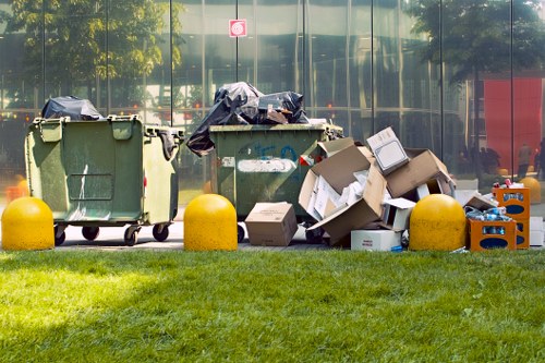 A row of three large wheeled waste bins in front of a glass building facade, with two of the bins covered by black trash bags and the third partially open, revealing some discarded cardboard and packaging materials. Next to the bins, there are several flattened cardboard boxes, some leaning against the bins and others lying flat on the grassy area. The scene appears to be an outdoor rubbish collection point, with the bins situated on a well-maintained grassy verge bordered by yellow protective bollards. The reflection of trees and a red structure can be seen in the glass behind, indicating the image was taken during daylight in a likely urban or suburban area, possibly near a commercial or residential property in Southend-on-Sea. The image represents waste collection and rubbish removal services in a typical outdoor setting, with clear details of materials and disposal items involved.
