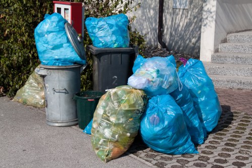 A collection of various rubbish bags and bins situated on a paved driveway near a residential building. The image shows multiple large blue plastic bags, some filled with waste and tied at the top, placed alongside grey and black wheelie bins, with one green bin partially visible. The blue bags appear to contain mixed household waste, including green and white items visible through the semi-transparent plastic. The grey bins are positioned closer to the building, while the black one stands slightly apart. Surrounding the area are some shrubbery and a section of concrete steps leading to a higher level, with part of a building wall visible in the background. The scene is lit naturally, suggesting daylight, and the setting appears to be in a suburban or residential area, consistent with local rubbish collection contexts such as those served by House Clearance Southend-on-Sea in the Southend-on-Sea postcode area.