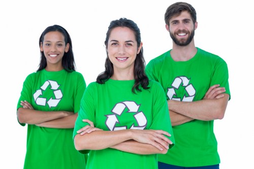 A group of three individuals standing outdoors on a paved surface with a building in the background, each wearing bright green t-shirts featuring a white recycling symbol. The woman in the middle has dark hair styled in loose waves and is smiling with her arms crossed. To her left, a woman with long dark hair is also smiling broadly, arms folded across her chest. To her right, a man with a beard and short dark hair is smiling and standing with his arms crossed. The scene appears to be a promotional image for waste management or recycling services, with the individuals representing environmentally friendly initiatives, aligned with rubbish removal companies like House Clearance Southend-on-Sea serving the local area of Southend-on-Sea, Essex.