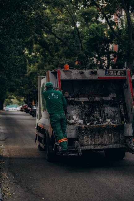 A waste collection worker wearing a green uniform, helmet, and orange reflective stripes is standing on the street beside a closed commercial rubbish collection vehicle, which is predominantly white w