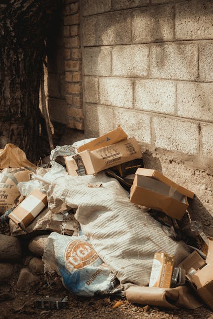 A waste collection worker wearing a green uniform, helmet, and orange reflective stripes is standing on the street beside a closed commercial rubbish collection vehicle, which is predominantly white w