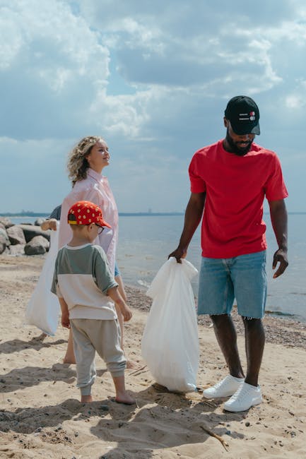 A man and two children on a sandy beach during daytime, participating in a rubbish cleanup activity near the water's edge. The man, dressed in a red T-shirt, light blue denim shorts, and a black cap,