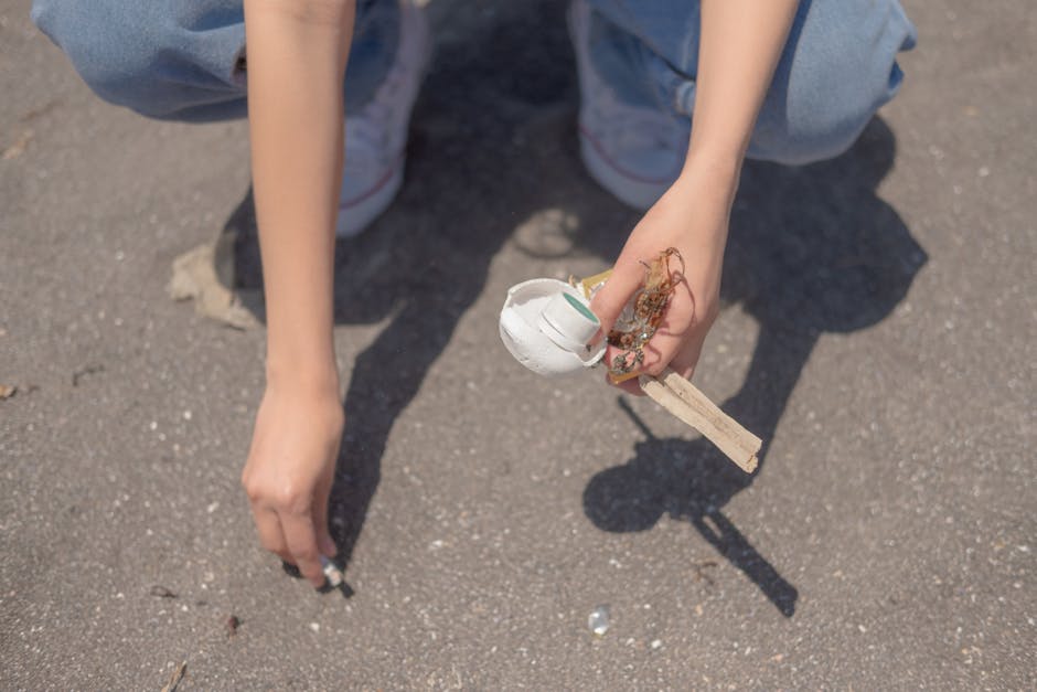 A man and two children on a sandy beach during daytime, participating in a rubbish cleanup activity near the water's edge. The man, dressed in a red T-shirt, light blue denim shorts, and a black cap,
