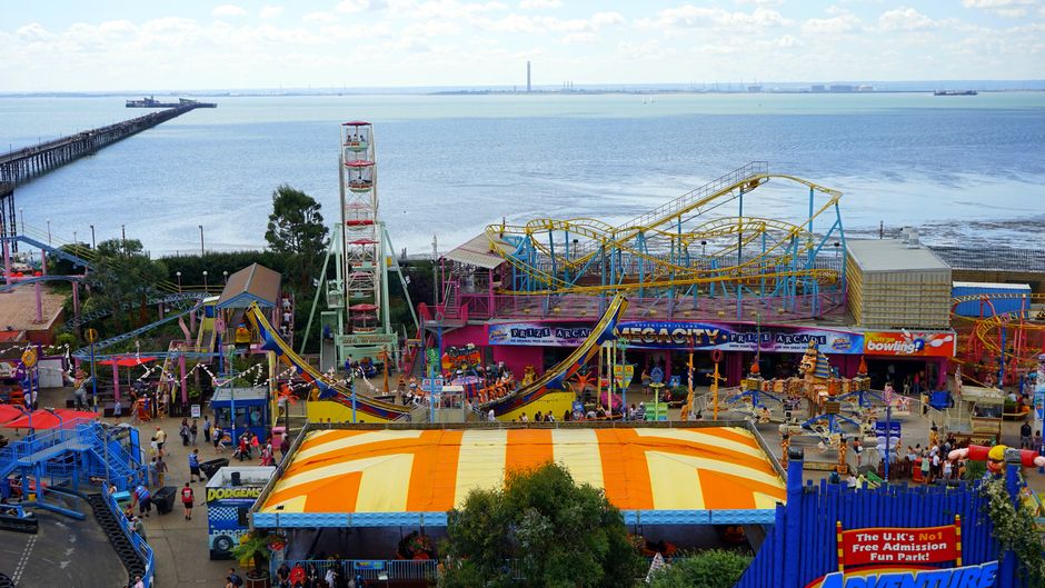 An aerial view of a busy seaside promenade during daylight, showing a long line of parked cars along the curb on the right side of the image, with some vehicles also occupying the street. On the left 